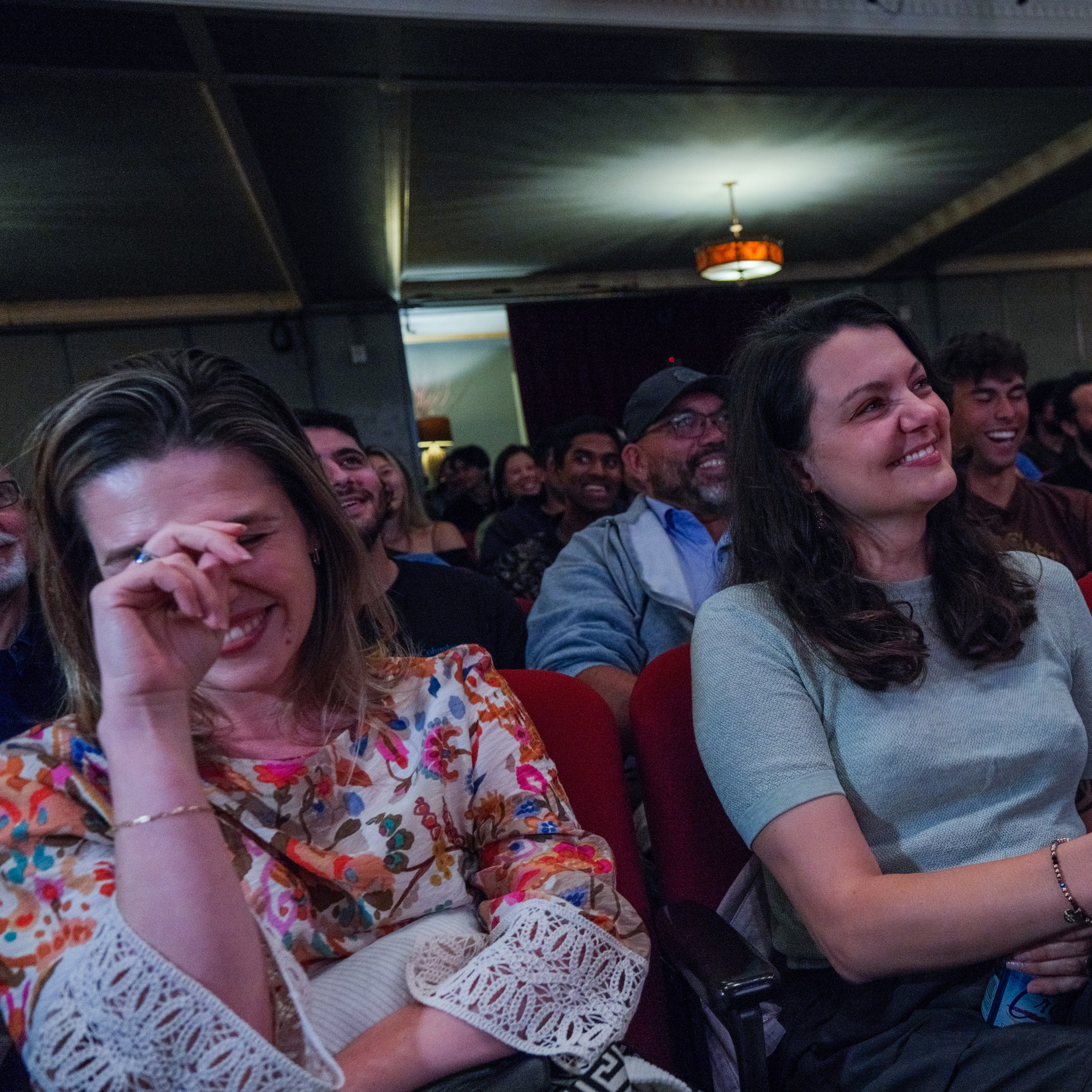 A crowd is enjoying a show, with two women in front; one covers her face, laughing, while the other smiles. The audience behind them is also laughing.