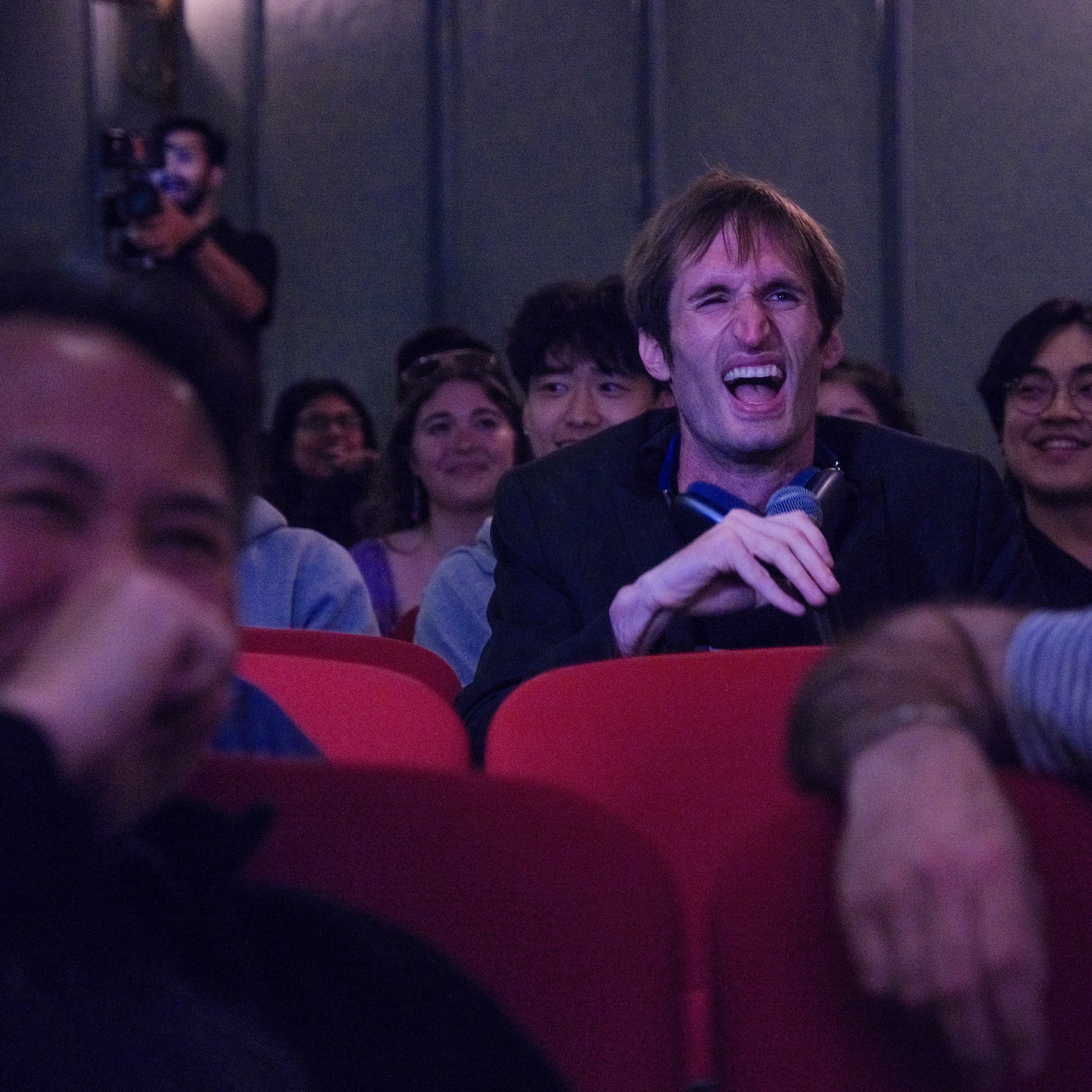 A man in a suit is laughing loudly while holding a microphone in a dimly lit theater. The audience around him is also smiling and laughing.