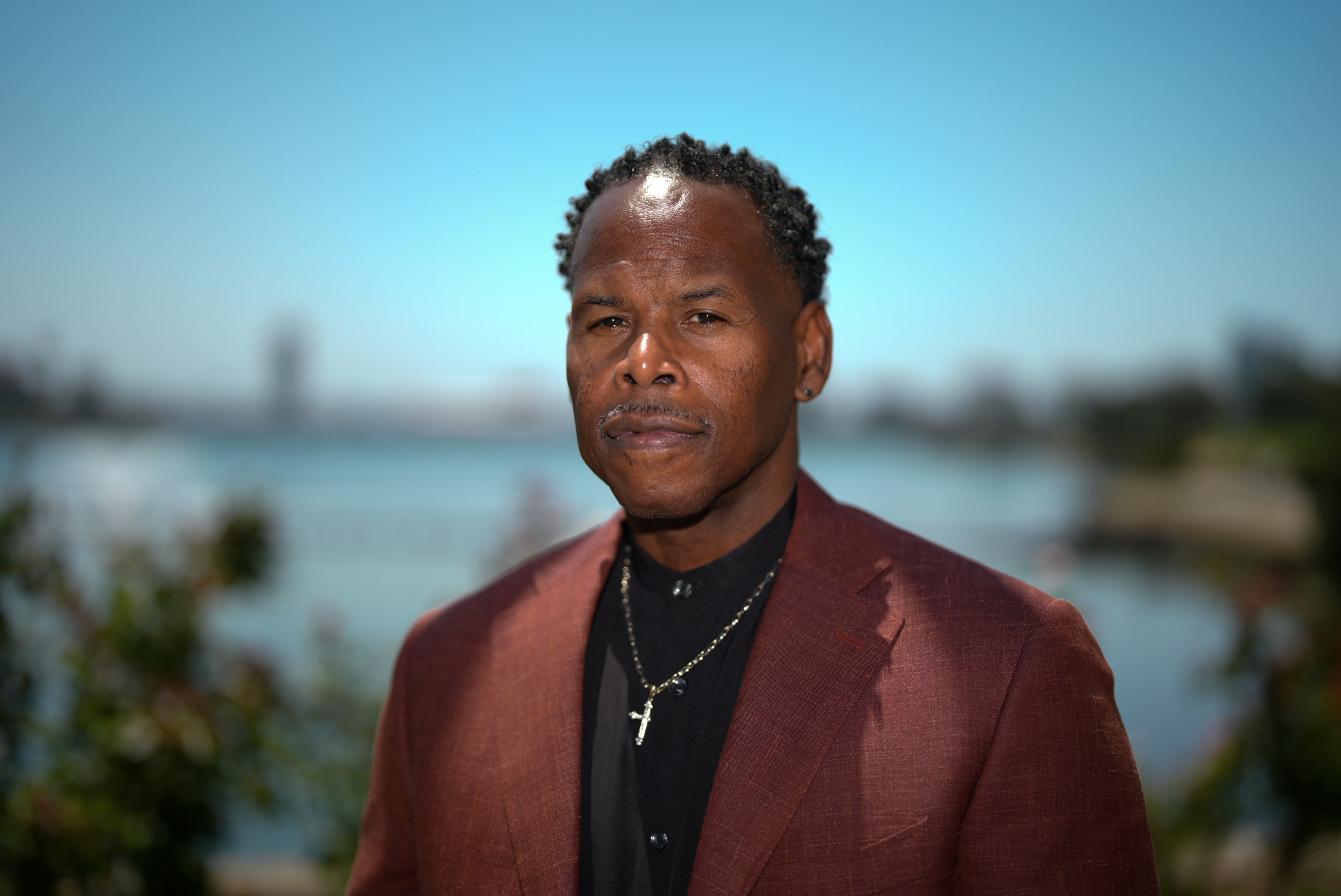 A man in a maroon blazer and black shirt stands outdoors against a blurred waterfront background, wearing a chain necklace with a cross pendant. The sky is clear and blue.