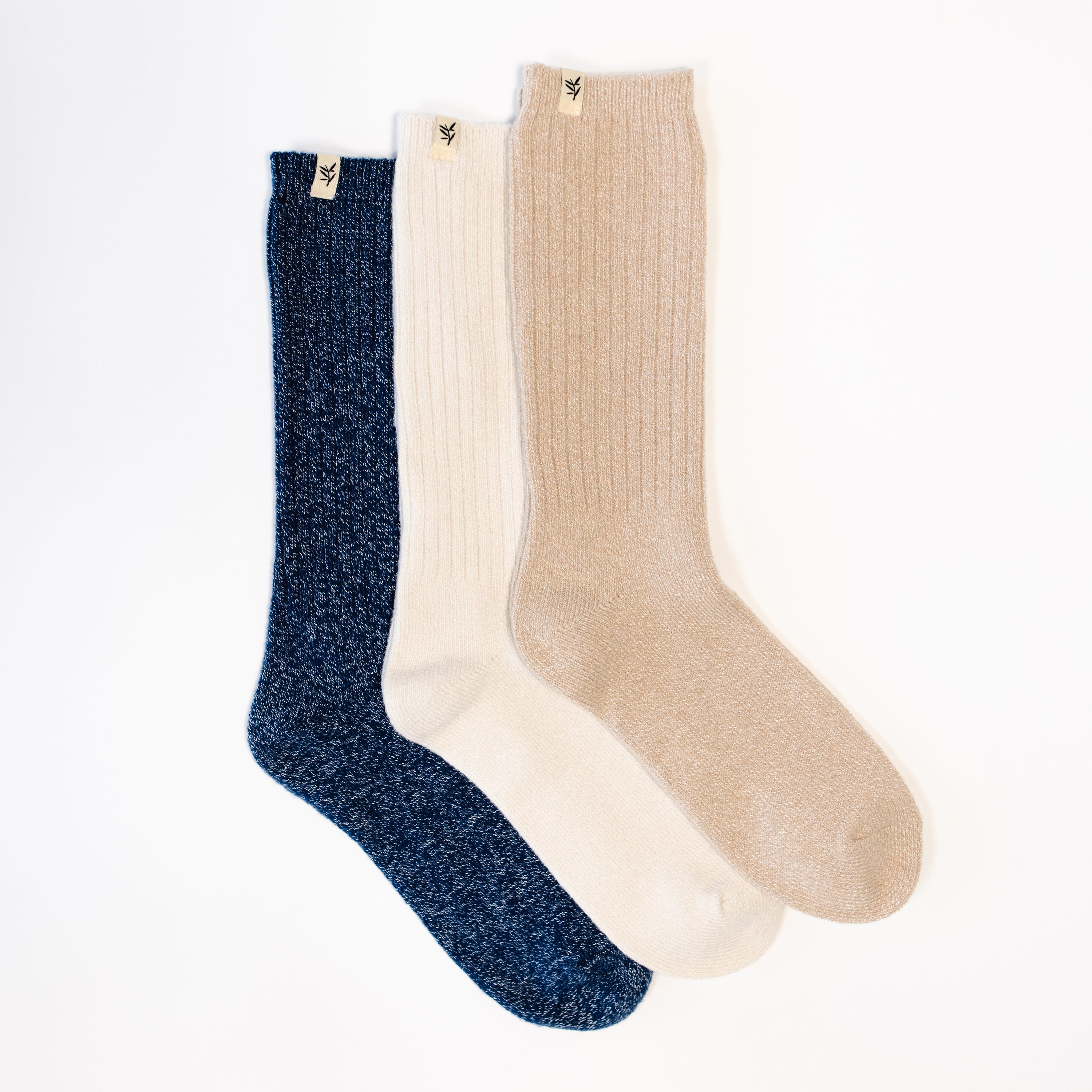 Three pairs of The Plush Lounge Sock by Cozy Earth are laid out on a white surface. The socks come in three different colors: Midnight Sky, Creme, and Almond. Each pair features a small tag with the logo at the top.