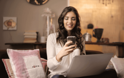 Mujer con cabello largo y vestido blanco en habitación sobre un sillón con celular en la mano y un computador sobre sus piernas