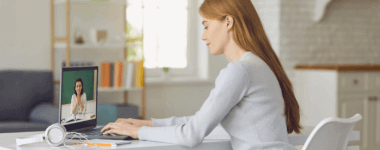 Mujer alegre frente a una pantalla de laptop recibiendo una clase por otra persona; la mujer esta sentada en una silla blanca y trabajando sobre una mesa desde una habitación