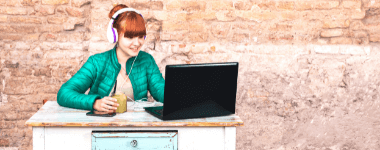 Mujer sonriendo mirando a la pantalla de una laptop con unos audiófonos en su cabeza; se encuentra sentada y los elementos en una mesa antigua y un fondo de pared de ladrillos