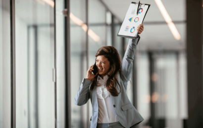 Mujer ejecutiva alegre hablando por telefono en una de sus manos y con la otra haciendo un gesto de victoria hacia arriba con un folder y algunas estadísticas en hojas de papel en ella; se encuentra en una oficina amplia y llena de ventanas