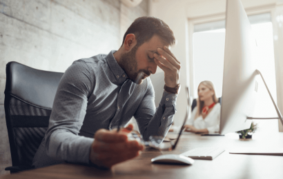 Hombre formal con ojos cerrados y su cabeza hacia abajo con una mano en su frente, trabajando en una oficina con un computador al frente de él