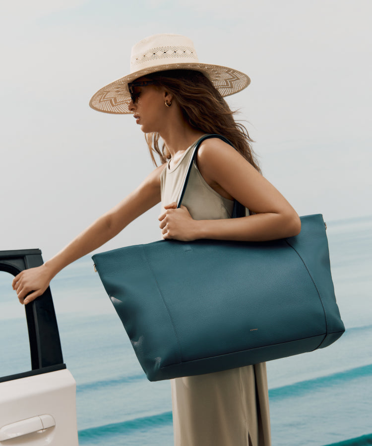 Woman in dress and hat carrying large tote bag near car by the beach.