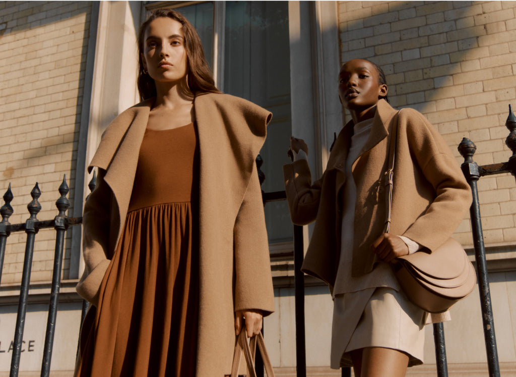 Two women standing in front of a building, both dressed in stylish outfits and holding bags.