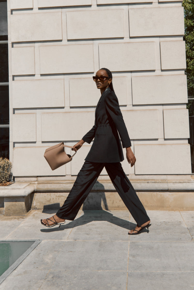 Woman walking past a wall carrying a bag and wearing sunglasses.