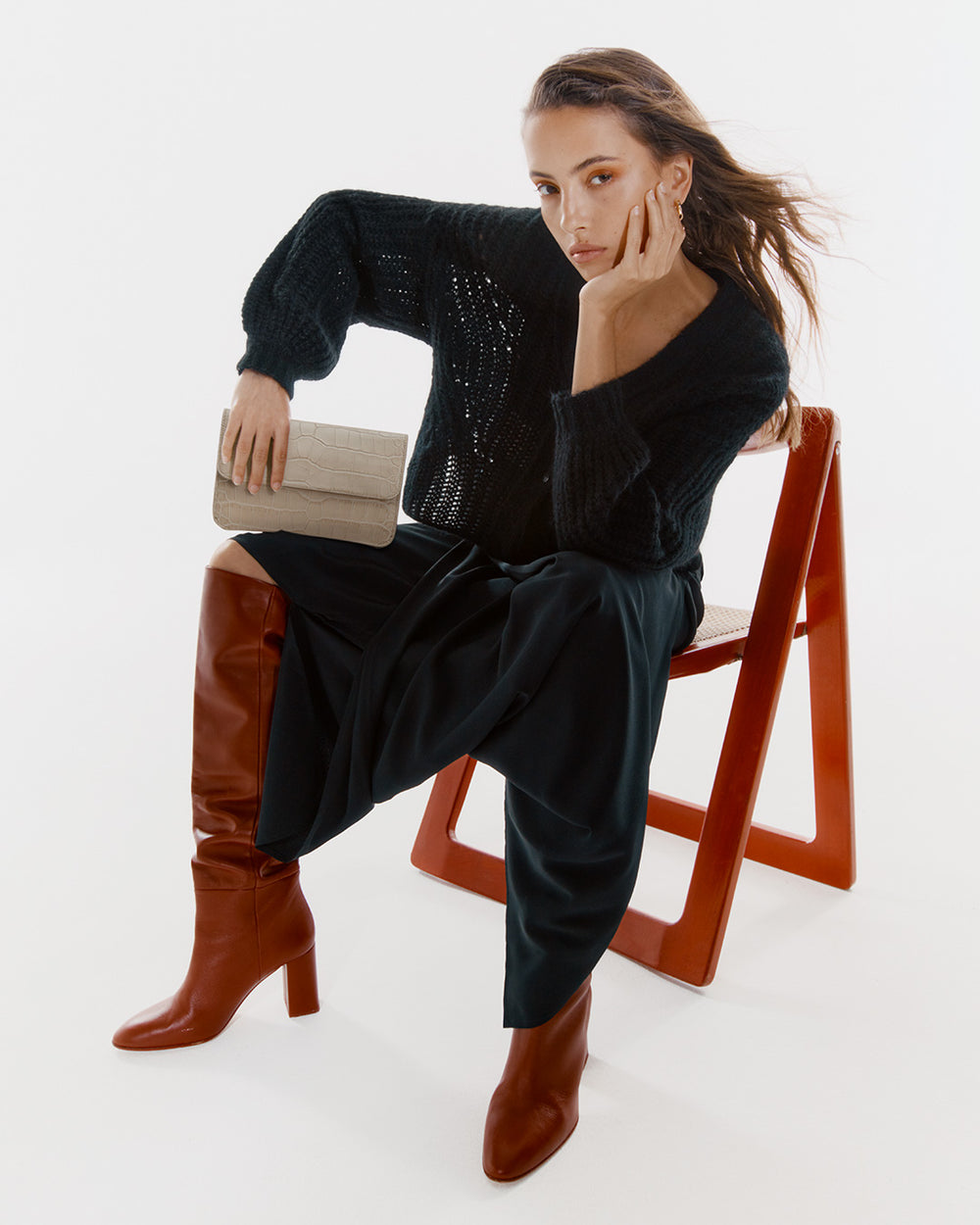 Person sitting on a chair holding a small clutch bag, wearing a long-sleeve top, skirt, and knee-high boots