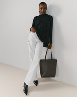 Person standing against a wall, dressed in a top and pants, holding a tote bag.