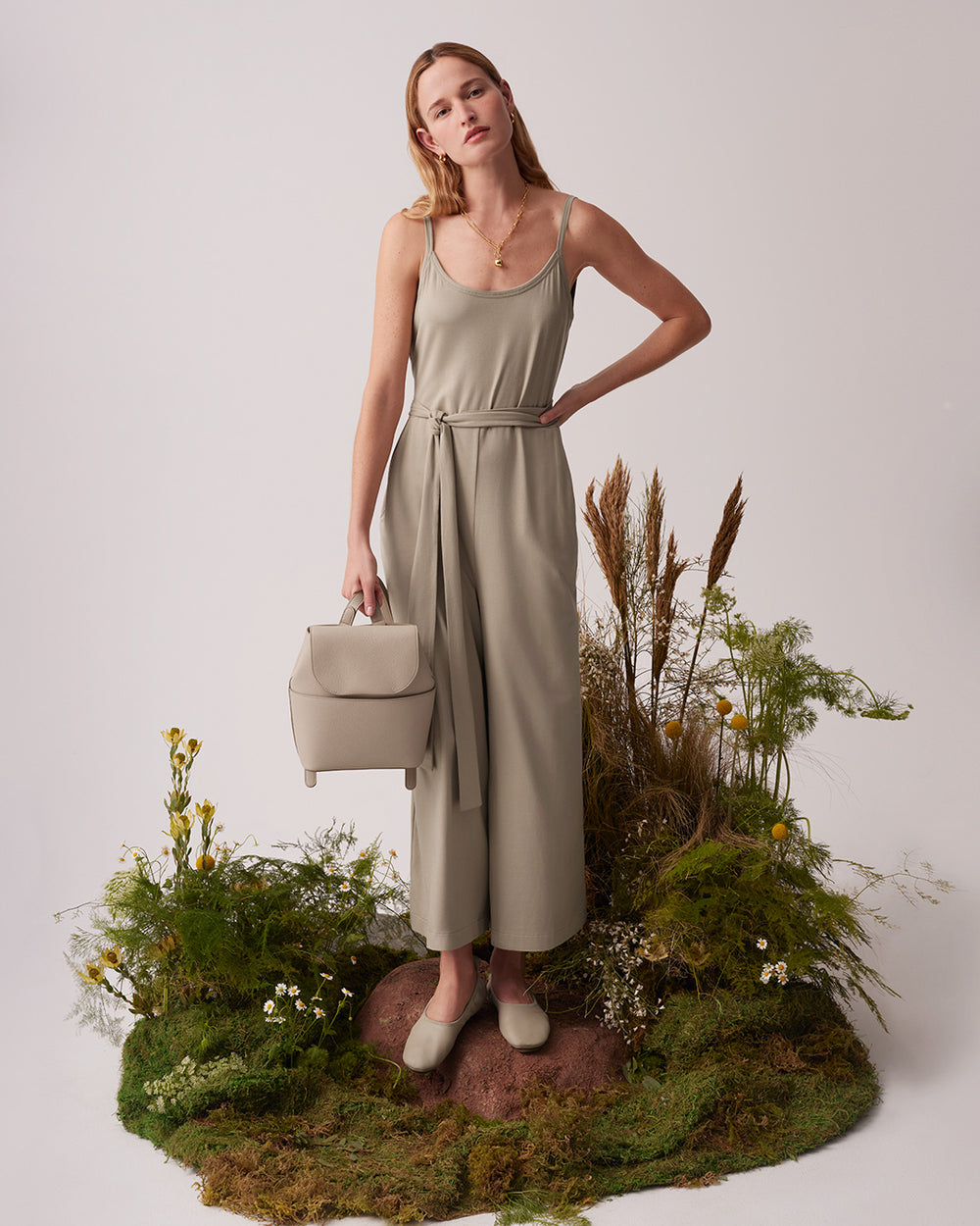 Woman in a dress standing on grass with a bag, surrounded by plants.