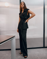 Woman standing in a room with hand on hip, wearing a sleeveless top and trousers.