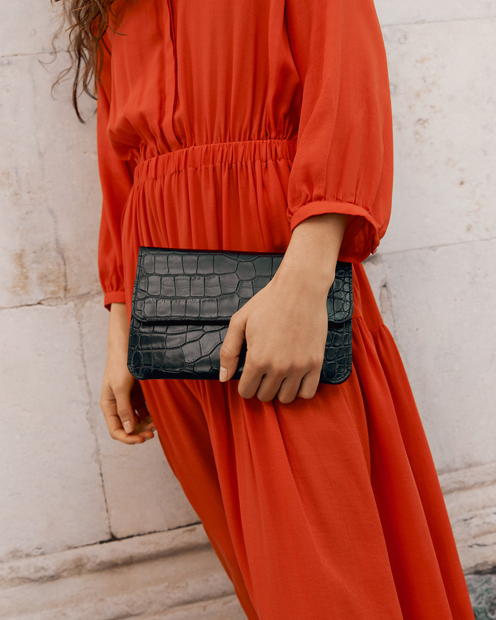 Person holding a croc-embossed leather clutch bag wearing a dress standing near a wall