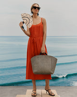 Woman in a dress holding a fan and a bag by the sea.