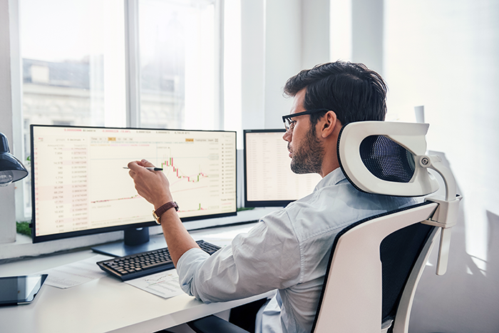 Analyzing data. Back view of young businessman or trader pointing on the data on computer screen with pen while working his modern office.