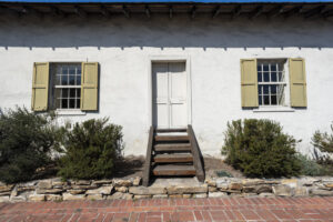 A white house with a white door in the center and wooden stairs leading up to it. On either side there is a white window with yellow shutters and a green shrub underneath.