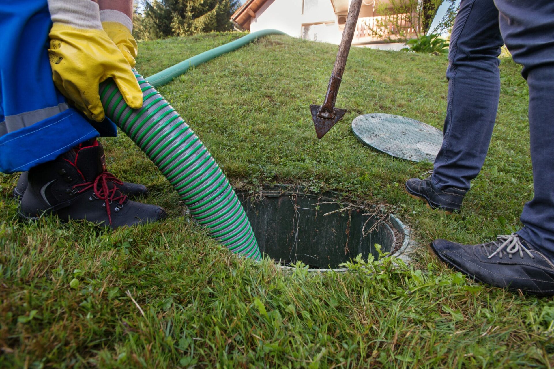 Two professionals with yellow gloves use a green hose to drain a septic tank in a green backyard.