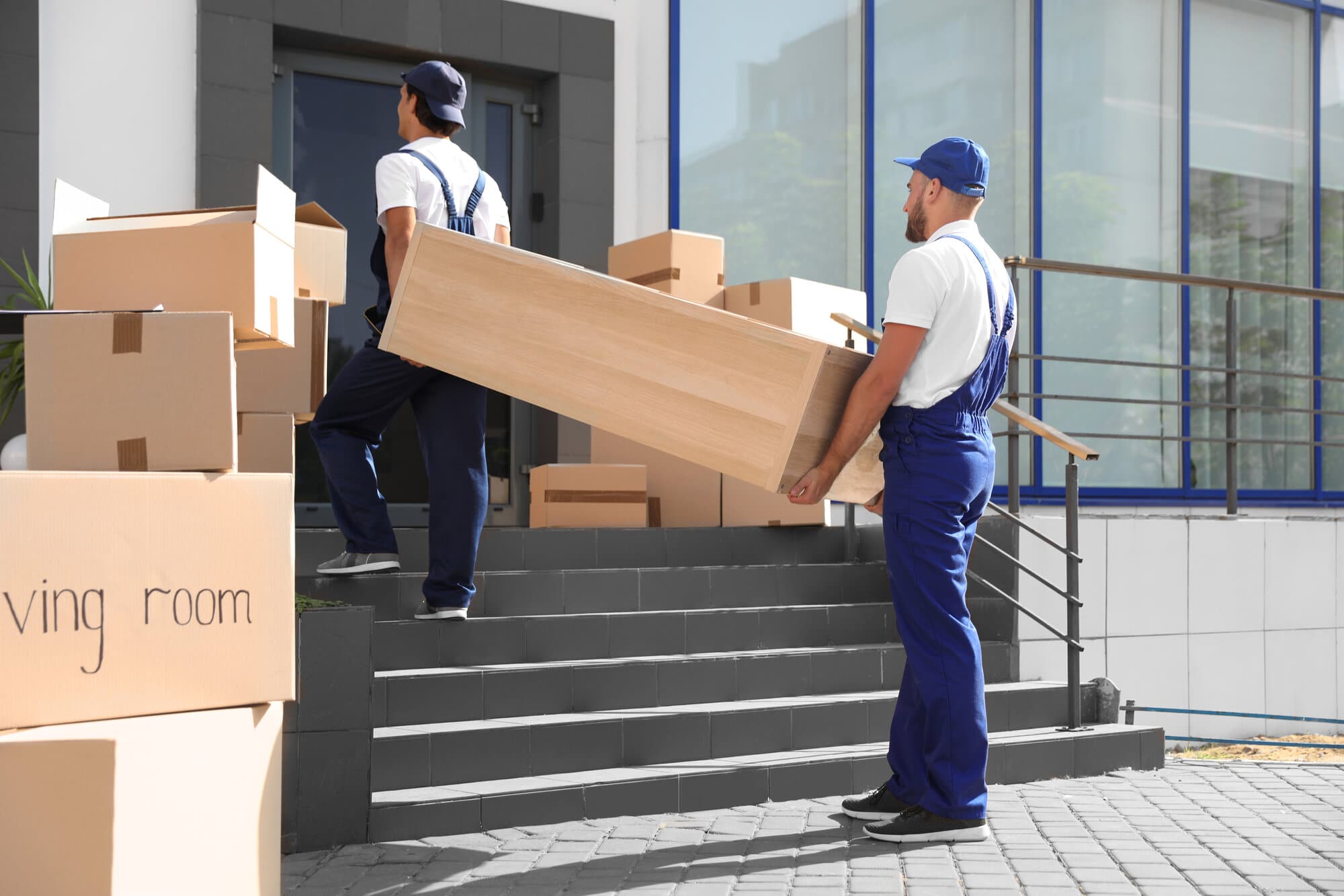 Male movers dressed in blue moving uniforms carrying boxes up the stairs into a house