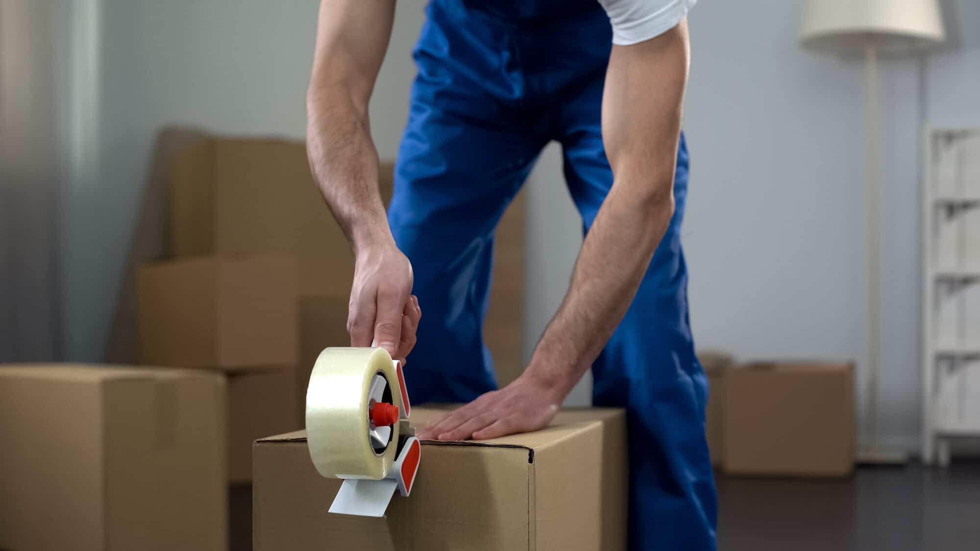 A closeup shot of a male mover using tape to tape up a cardboard box with more boxes in the background