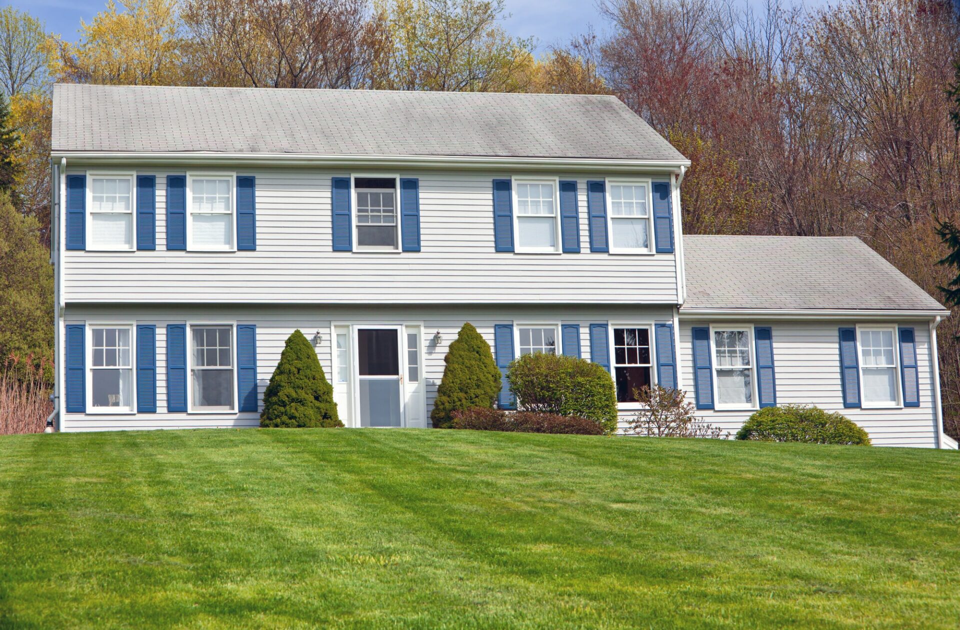 Colonial style White House with blue window shutters on a grass lawn with fall colored trees in the background