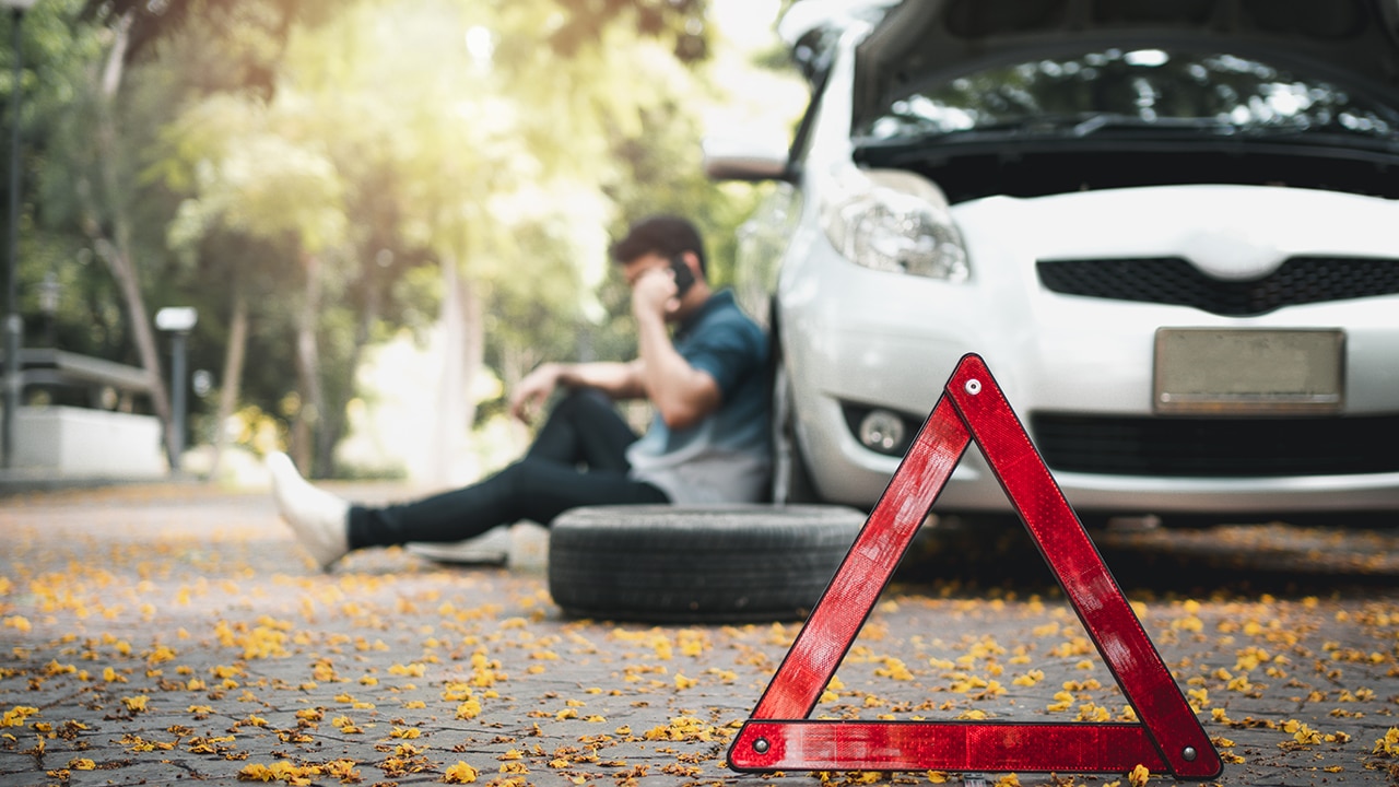 Asian man sitting beside car and using mobile phone calling for assistance after a car breakdown on street.