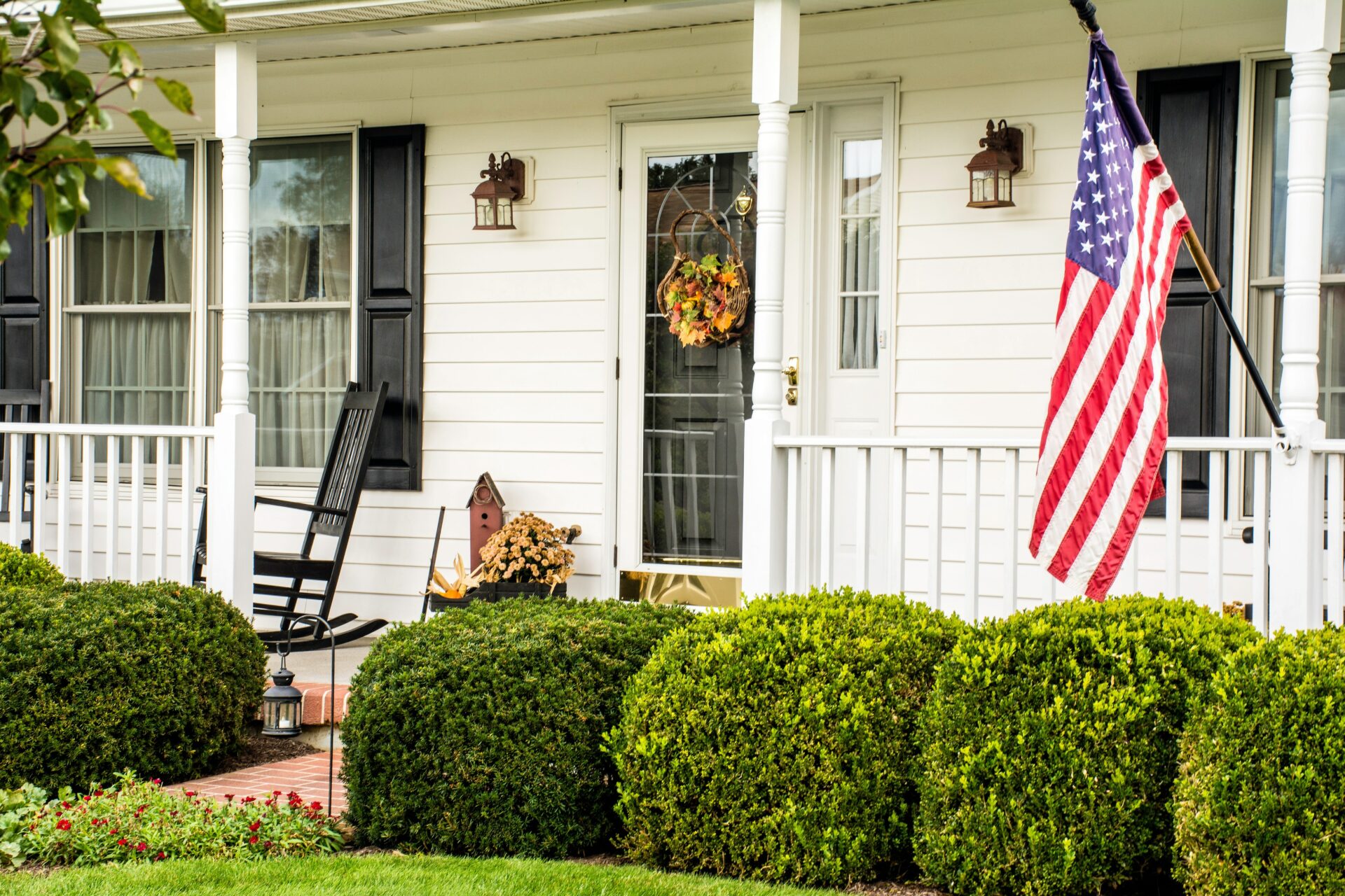 White colonial home with an American flag flying from the front porch with rocking chairs.