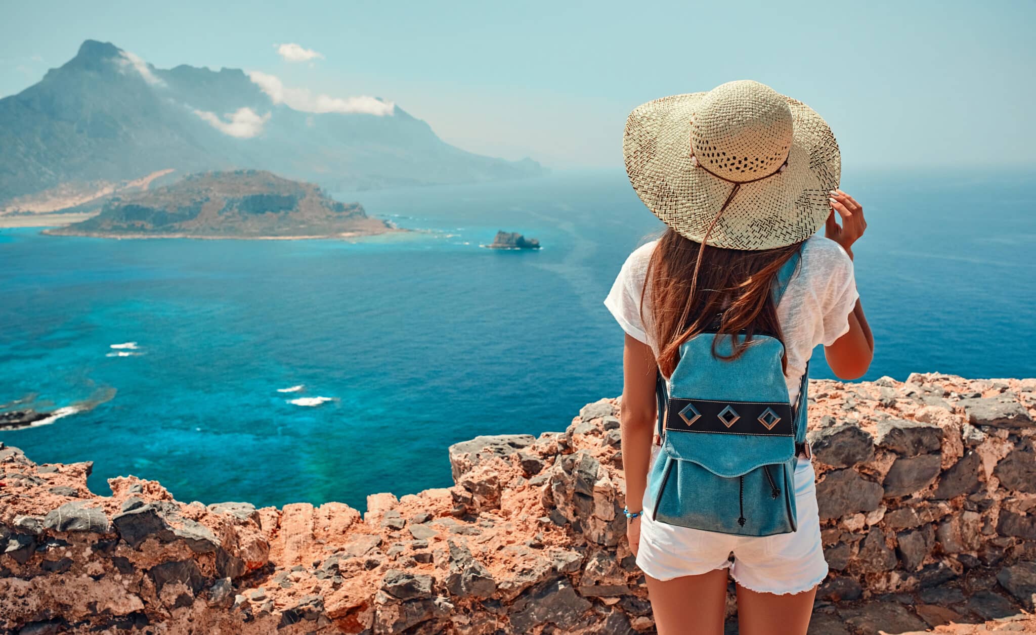 Portrait of happy girl in a hat with a backpack standing on background of mountains and sea.