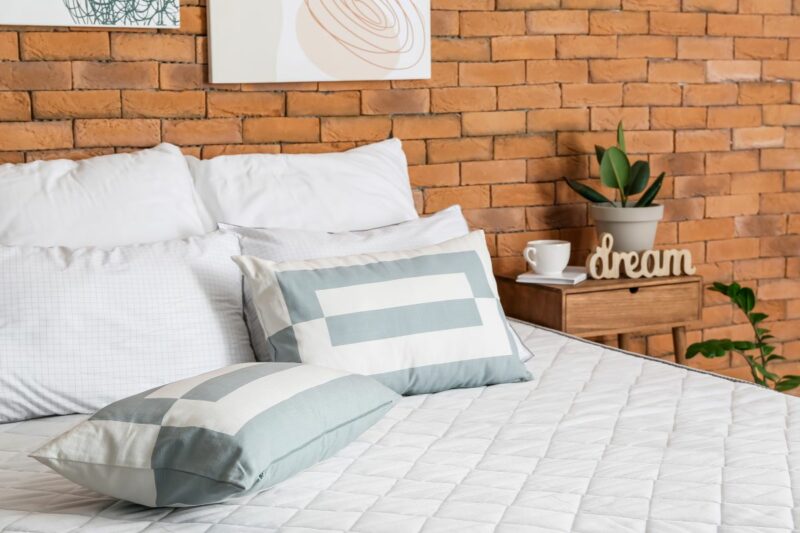 Bed with white quilted cover and white pillows in room with red brick wall and side table with coffee cup, plant, and carved word art spelling the word dream