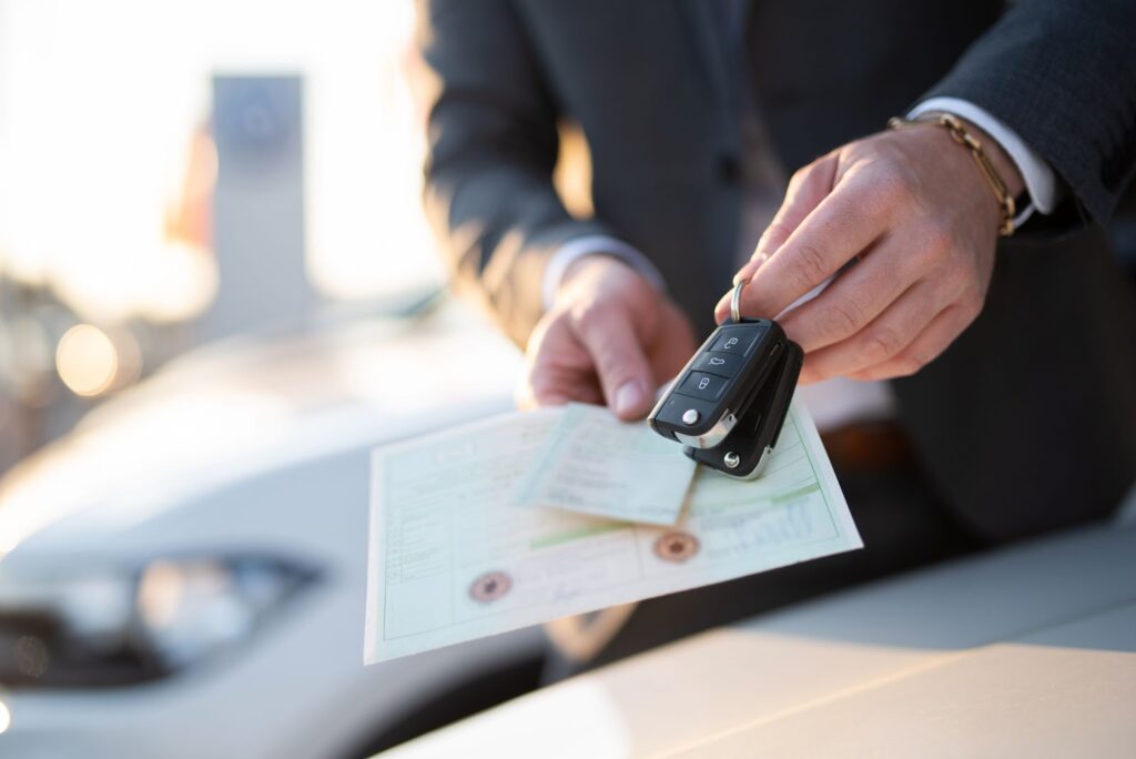 A person holds out a key fob and some documents with cars visible in the background