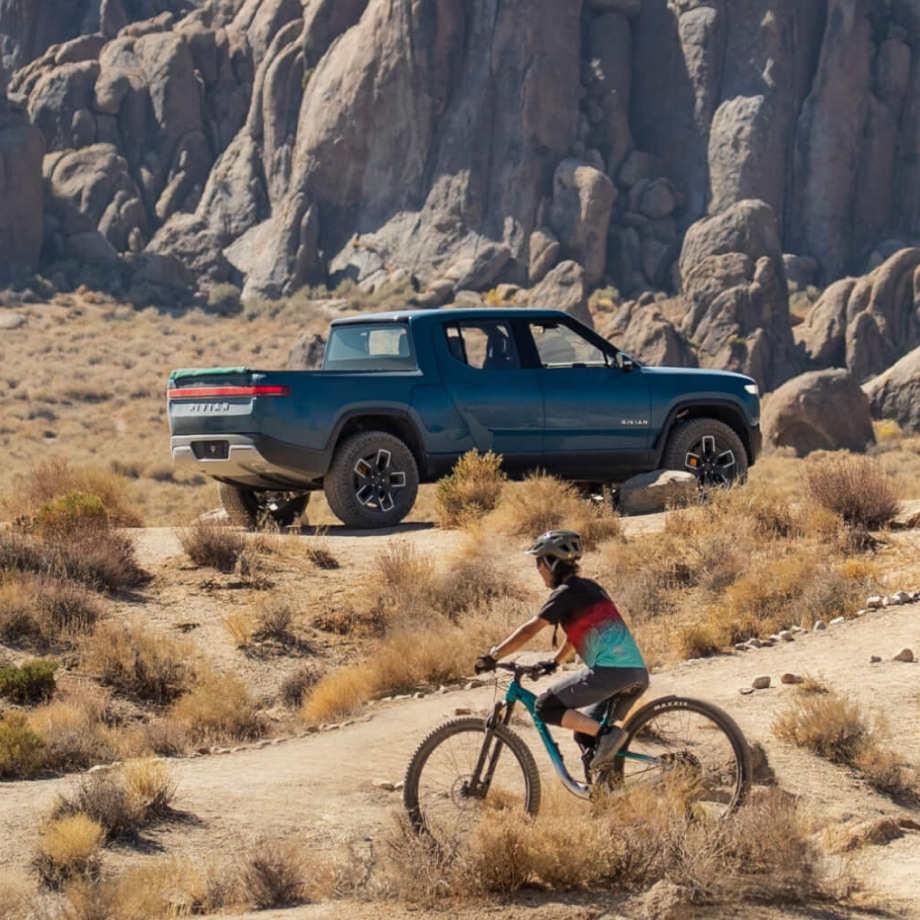 Rivian vehicle in a rugged landscape with bikers biking in front