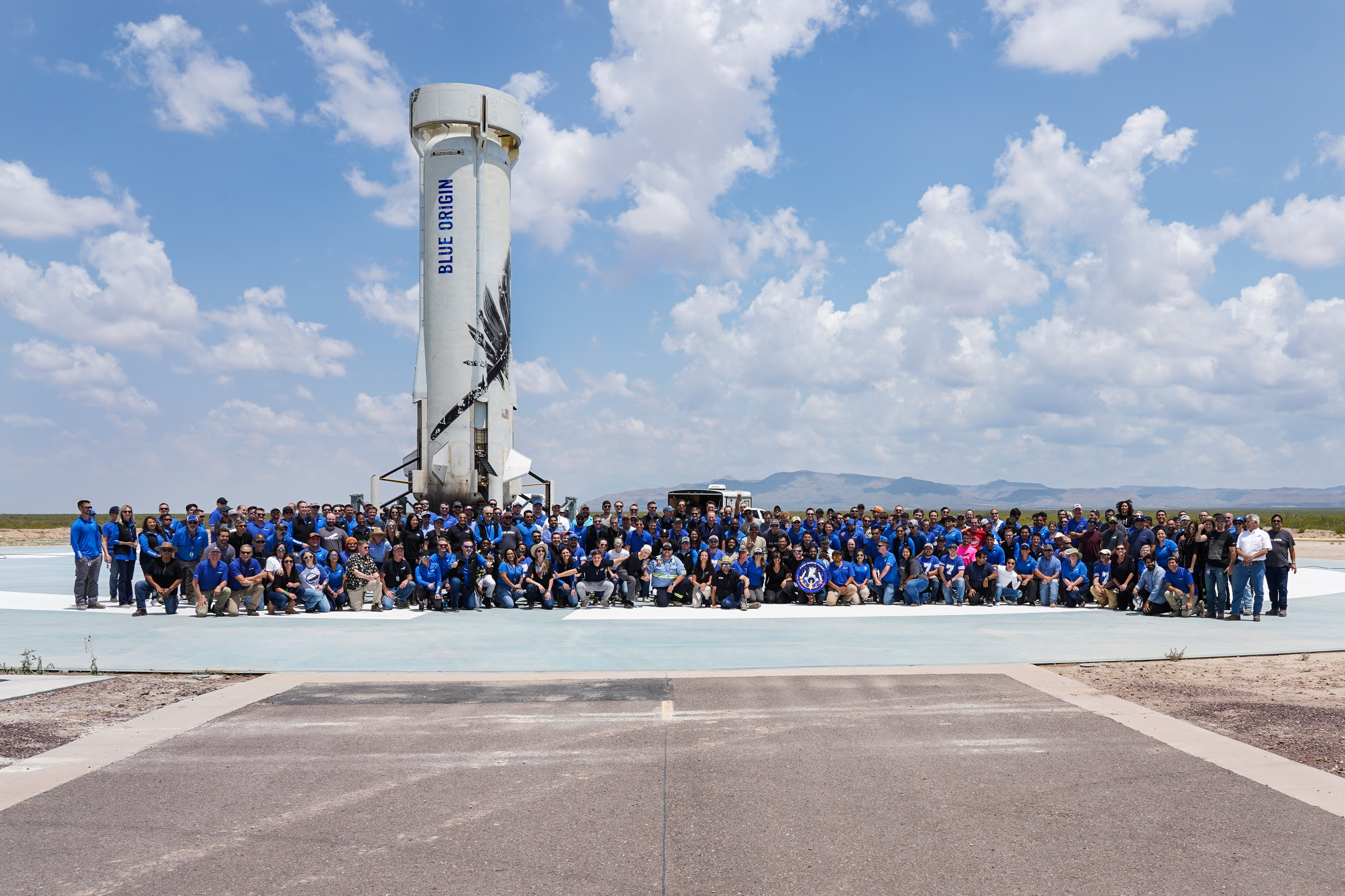 Hundreds of Blue Origin employees stand and kneel in multiple rows on the landing pad in front of the booster that powered New Shepard's first four crew members to space. An employee in the center of the first row holds a sign emblazoned with the First Human Flight mission patch; white billowy clouds fill the blue sky.