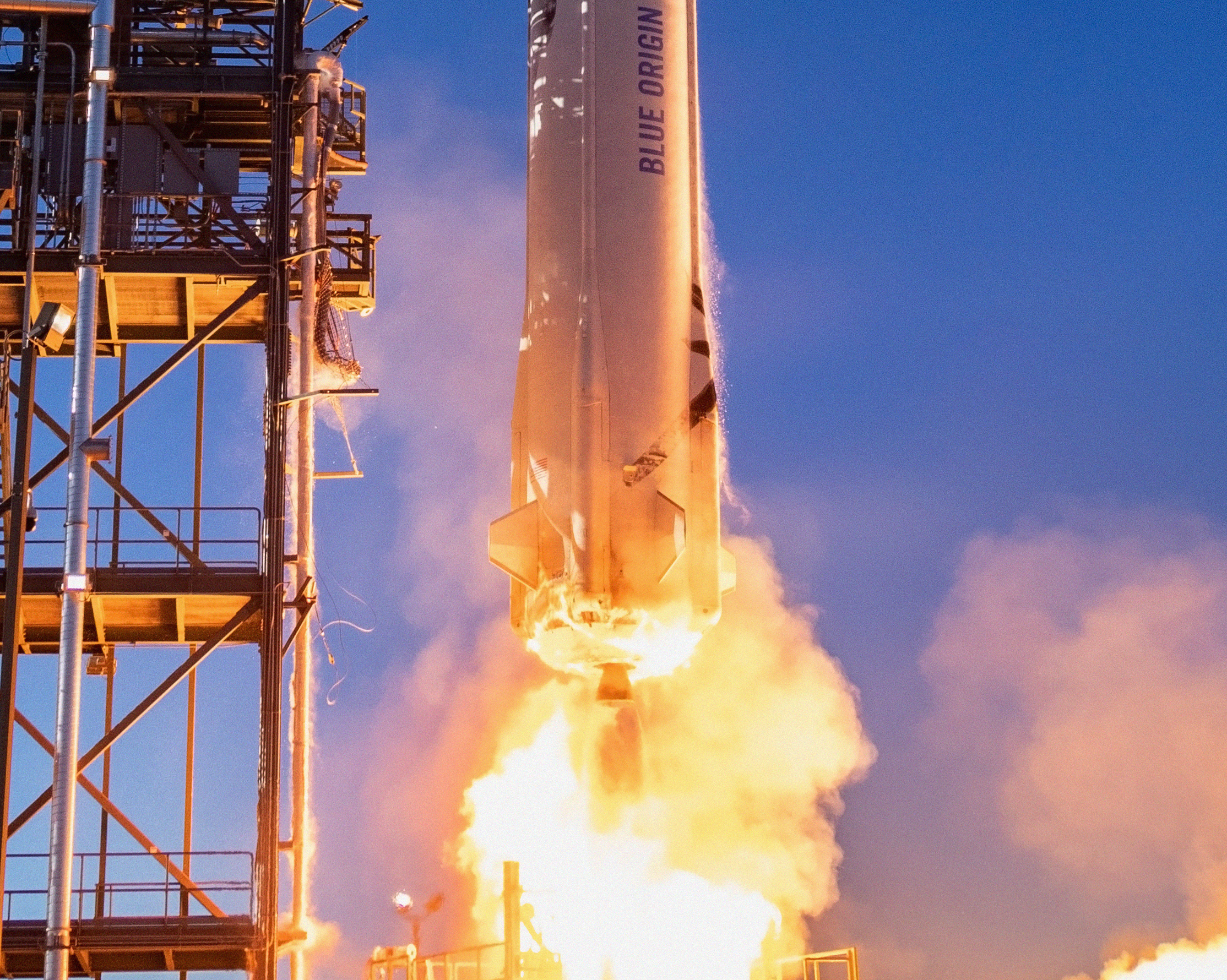A close-up of the bottom half of New Shepard, including the engine nozzle, next to the launch tower just after liftoff. A plume of fiery exhaust and light pink smoke clouds surround the vehicle.