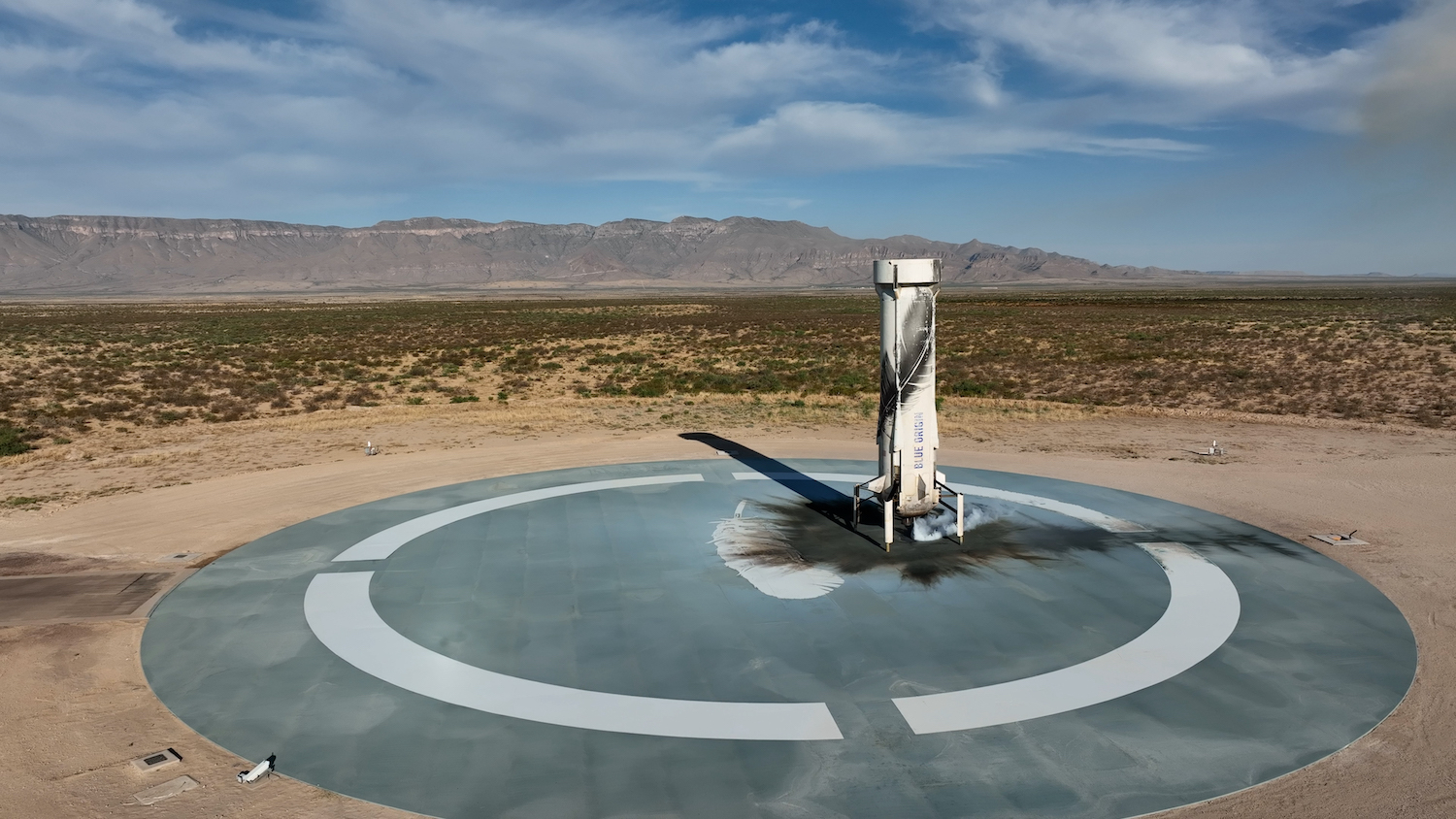 The booster is vertical on the landing pad. The area on the pad immediately around the booster is scorched by the engine's flames, and the desert and mountains can be seen in the background.