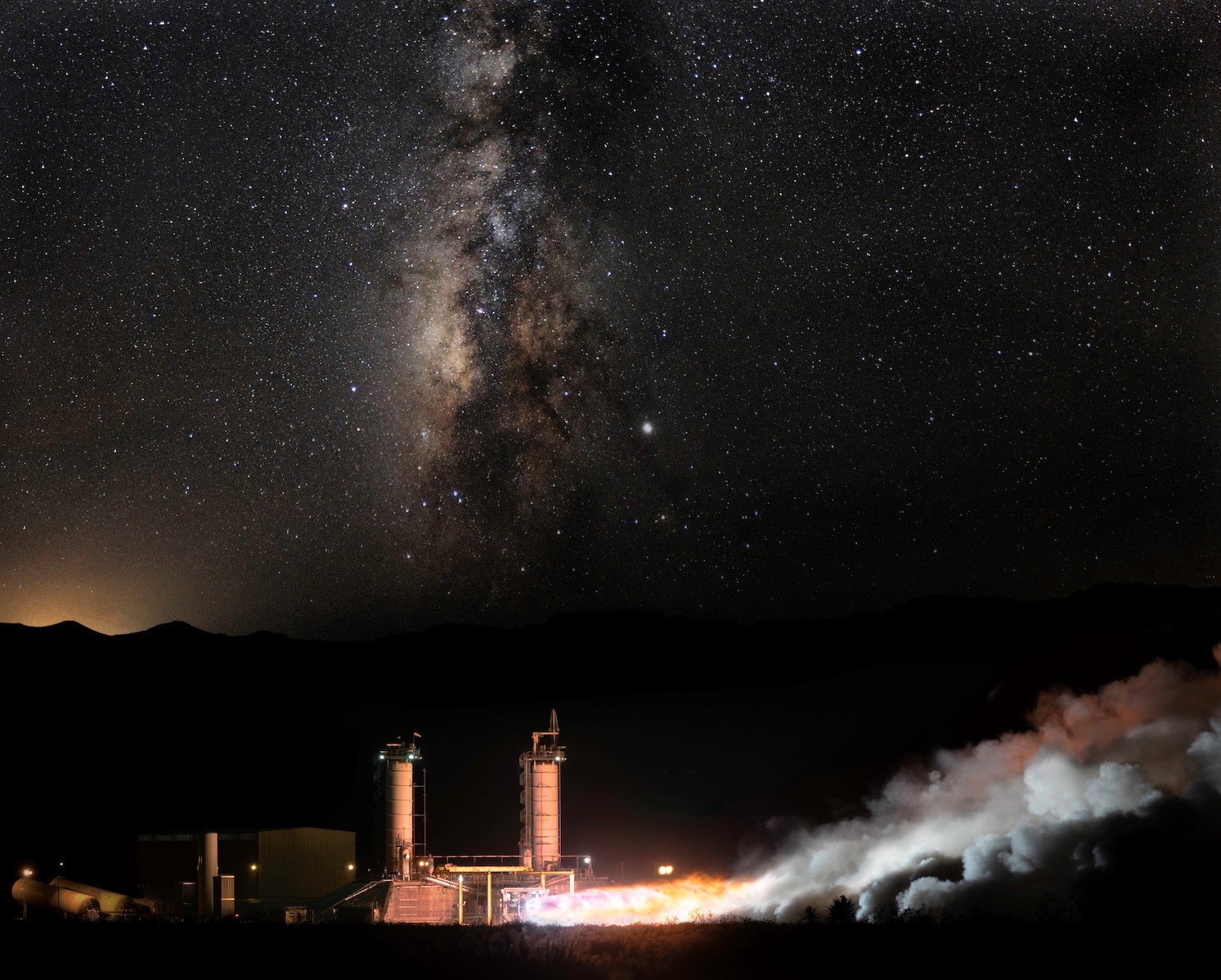 The BE-4 is hotfired on the test stand, expelling fiery exhaust that transforms into horizontal plumes of white smoke. The sky is filled with bright stars, including what appears to be a nebula in the sky above the test stand.