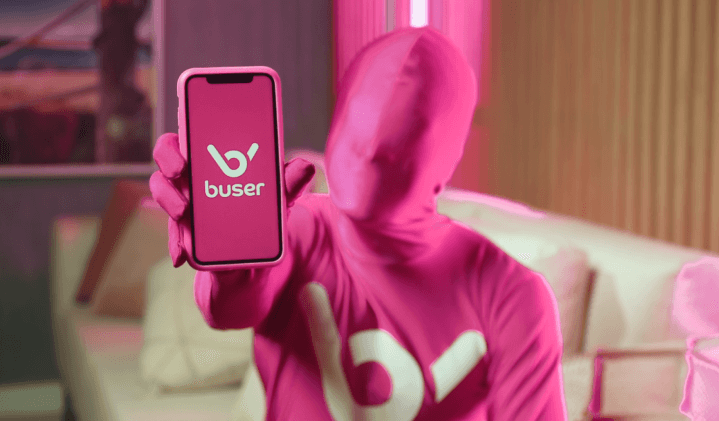 Imagem do boneco rosa da Buser apontando a tela do celular em direção ao usuário da plataforma