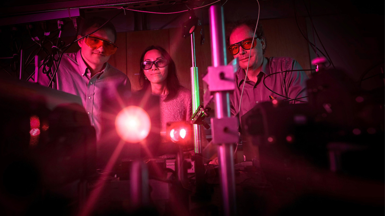 Two students and a professor stand around a table with a brightly glowing red beam 