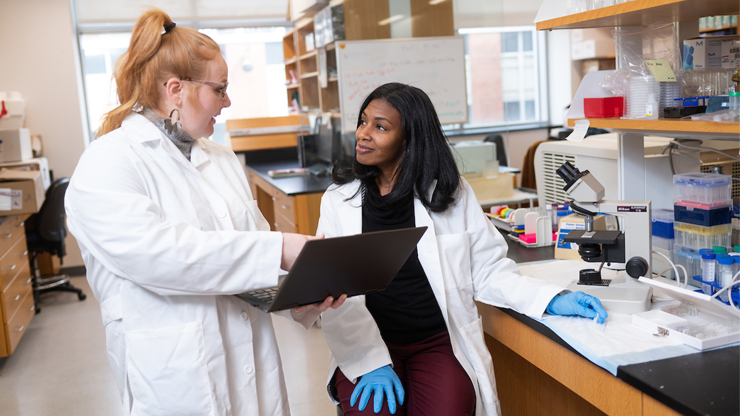 Two women wearing white lab coats talk in a laboratory while looking at a laptop screen