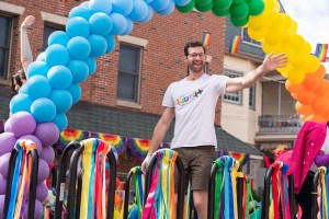 A screencap from 'Bros' in which Billy Eichner is standing on a Pride Parade float. Above him is an arch of rainbow balloons, and below him are an assortment of rainbow streamers lining the float.
