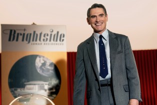 Billy Crudup standing in front of a poster that says "Brightside" in 'Hello Tomorrow!' on Apple TV+