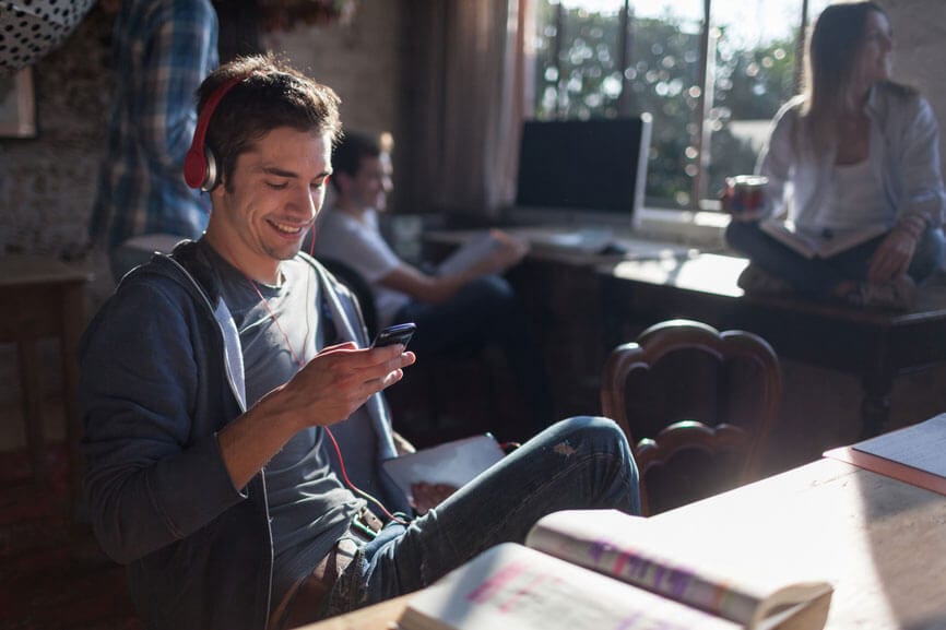 A young man wearing headphones and seated in a chair smiles and looks at his cell phone.