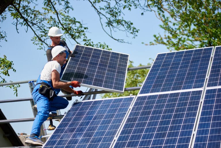 Two workers installing heavy solar photo voltaic panel on tall steel platform on green tree and blue sky background. Exterior stand-alone solar panel system installation.