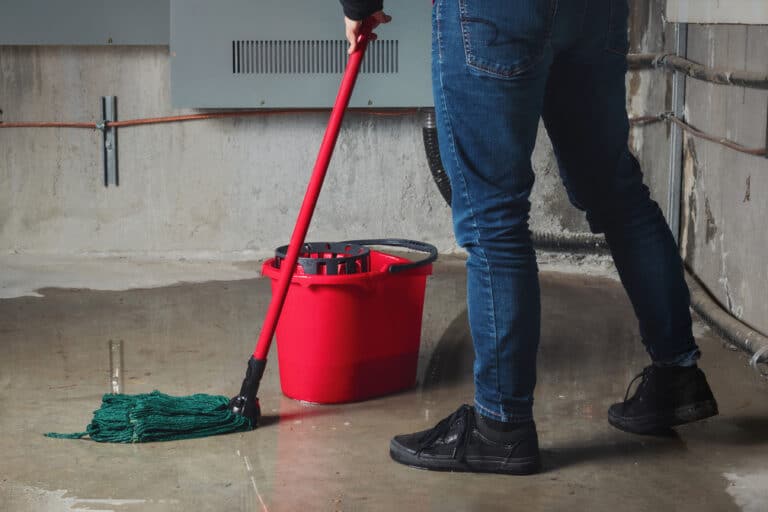man with a red bucket and mop cleans up water on concrete floor