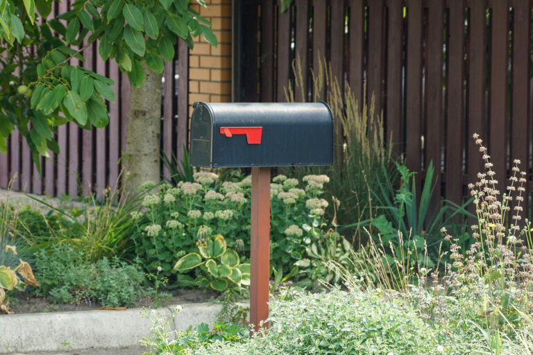 Black mailbox with a red flag at the end of a driveway. Greenery behind mailbox.