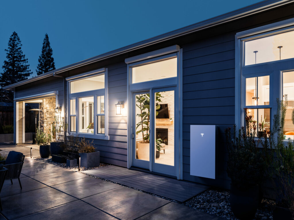 Modern house at nighttime with large windows. White Tesla Powerwall next to the back door.