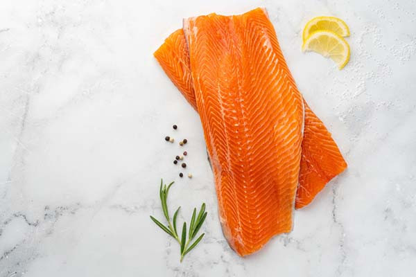 photo of fresh uncooked lake trout fillets on a table top alongside lemon slices, peppercrons, and rosemary leaves