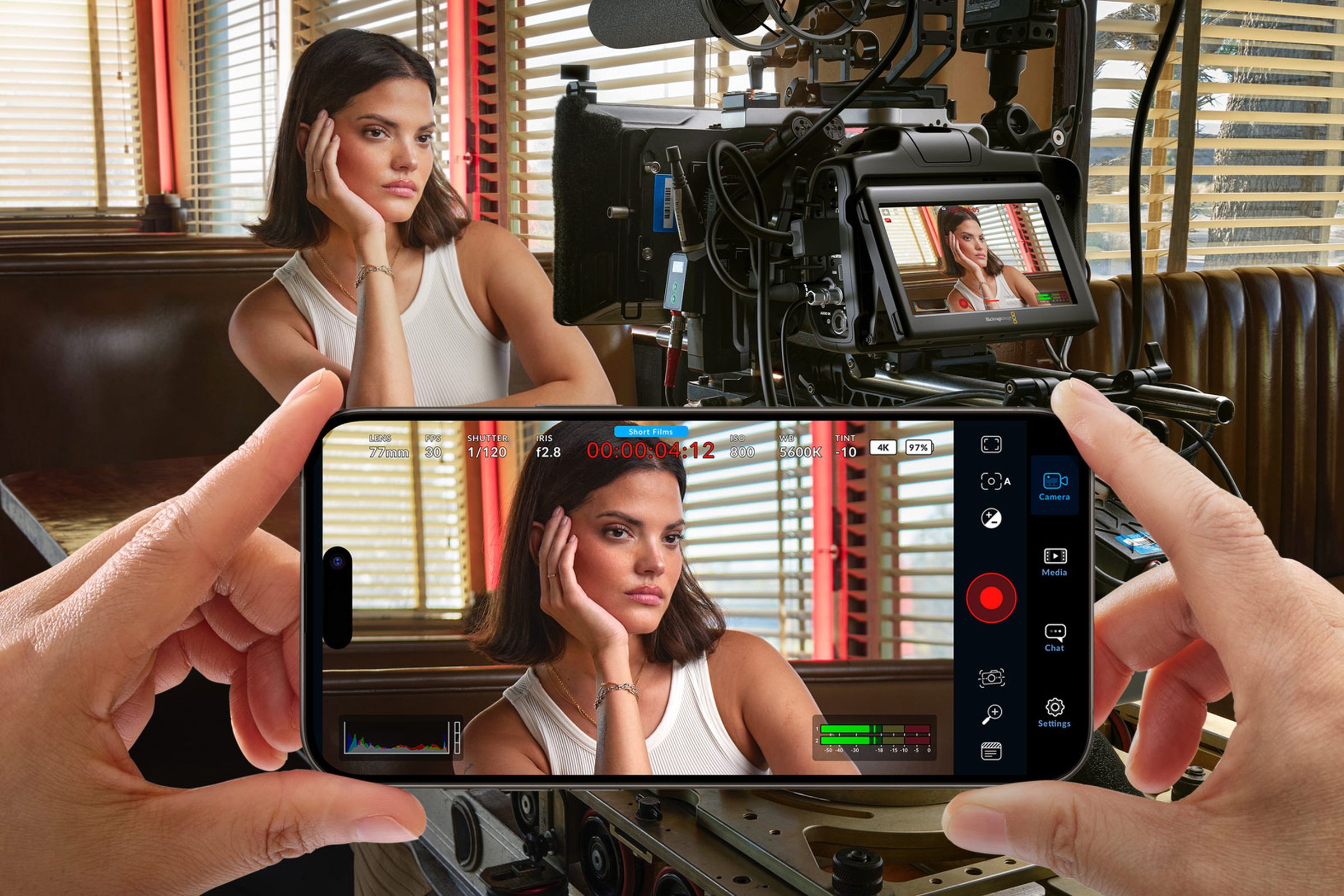 An actor in a diner is framed using the Blackmagic Camera app on a smartphone, behind a larger professional-grade camera.