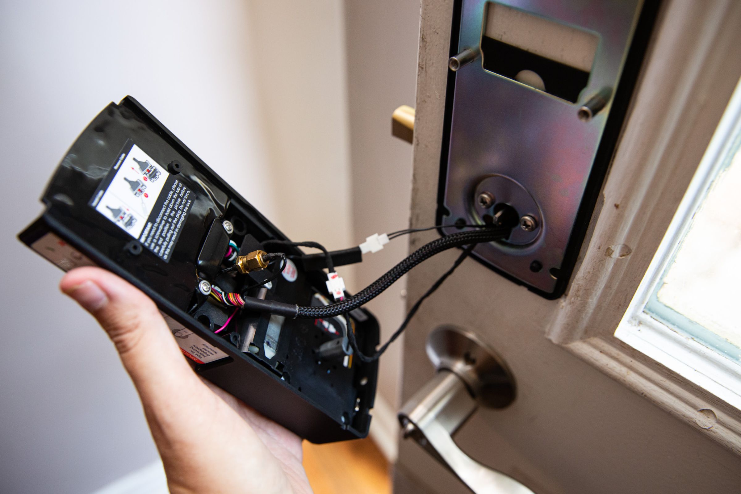 Hand holding the interior side of the Lockly Vision Elite door lock, detached from the door but connected via several cables through the deadbolt mechanism.