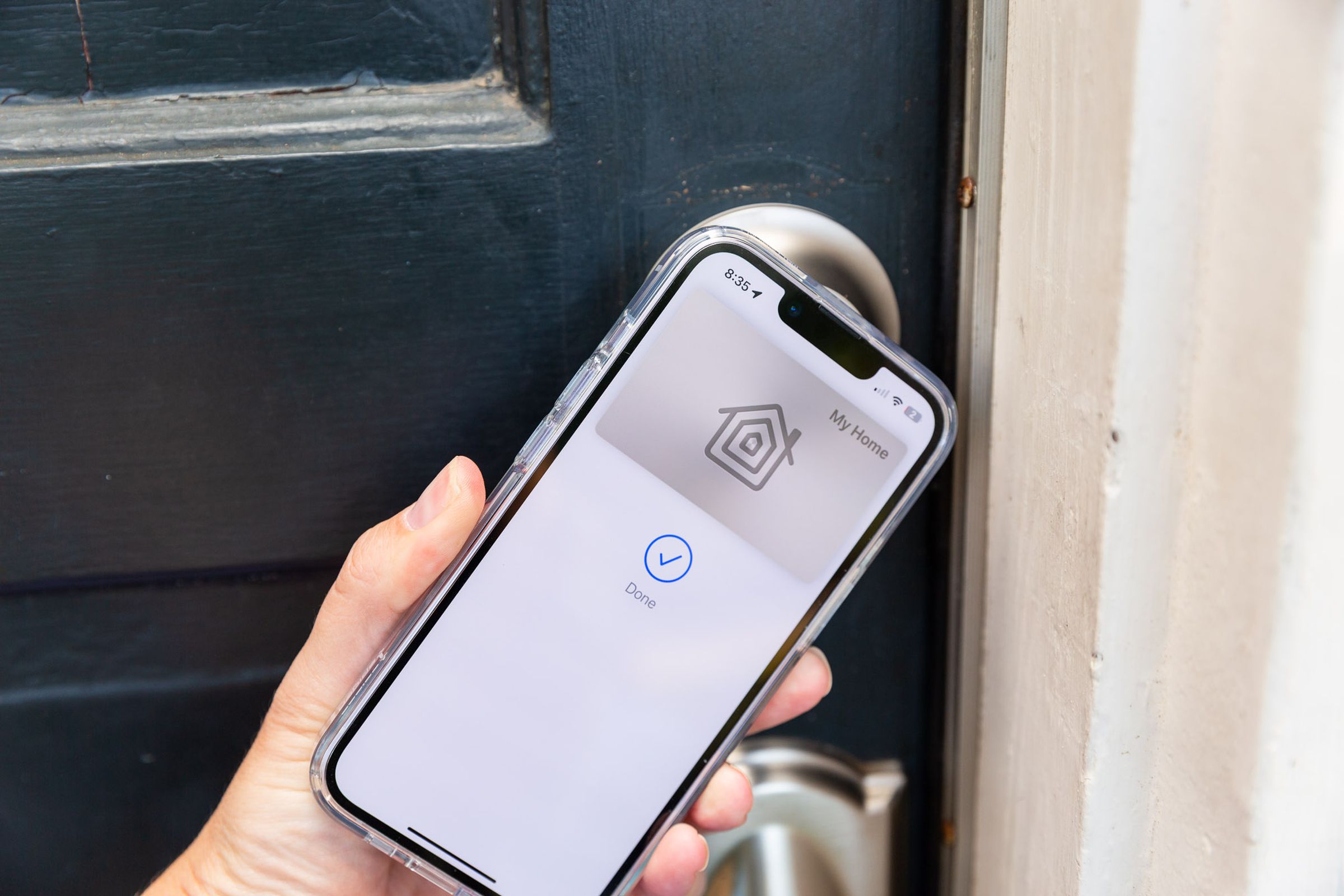 A hand holding an iPhone up to a door. On the phone screen is a picture of a keycard with the Apple Home logo, and the words “My Home” above it. Below it is a blue checkmark in a circle and the word “Done.”