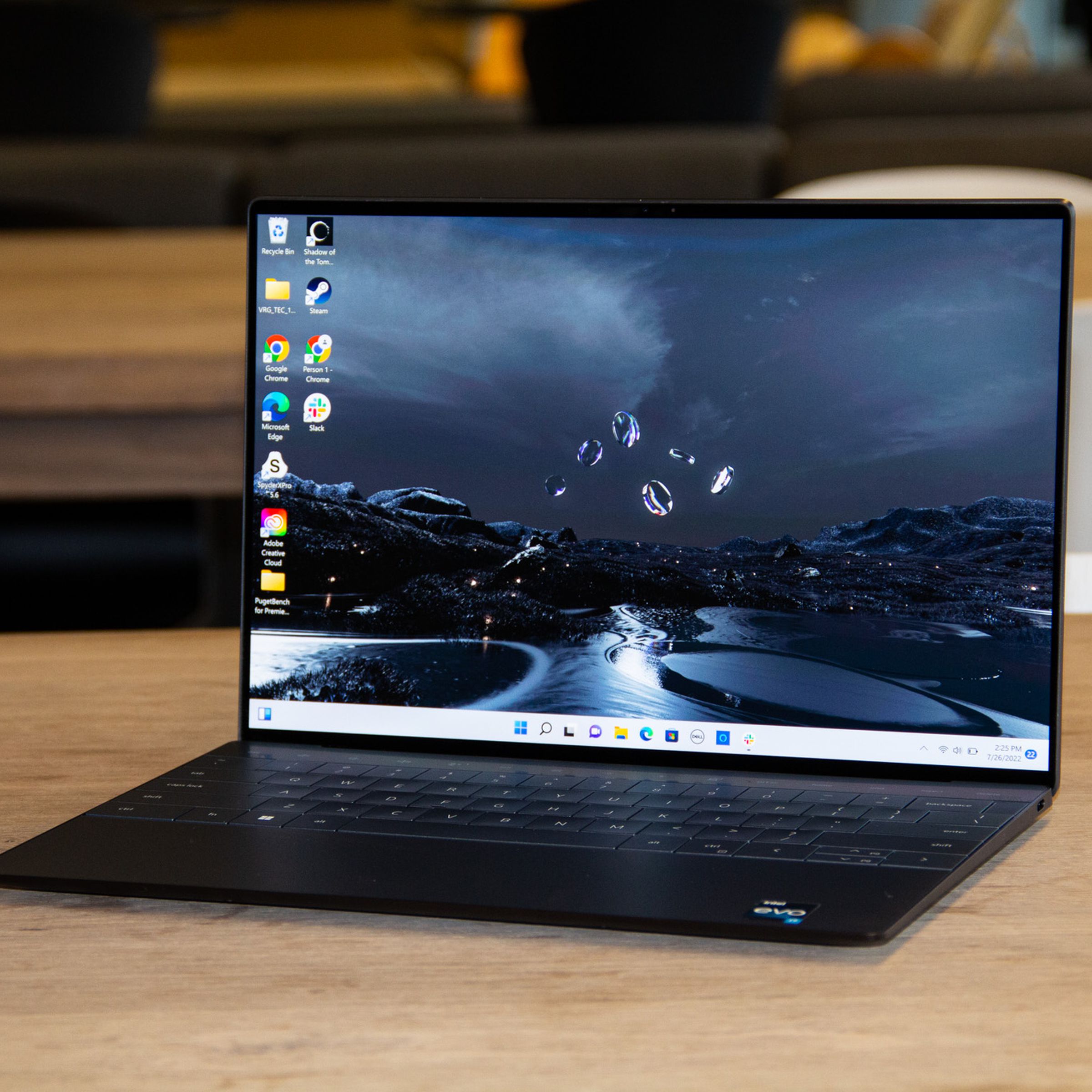 The Dell XPS 13 Plus open on a wooden table. The screen displays a river at night as a Windows desktop background.