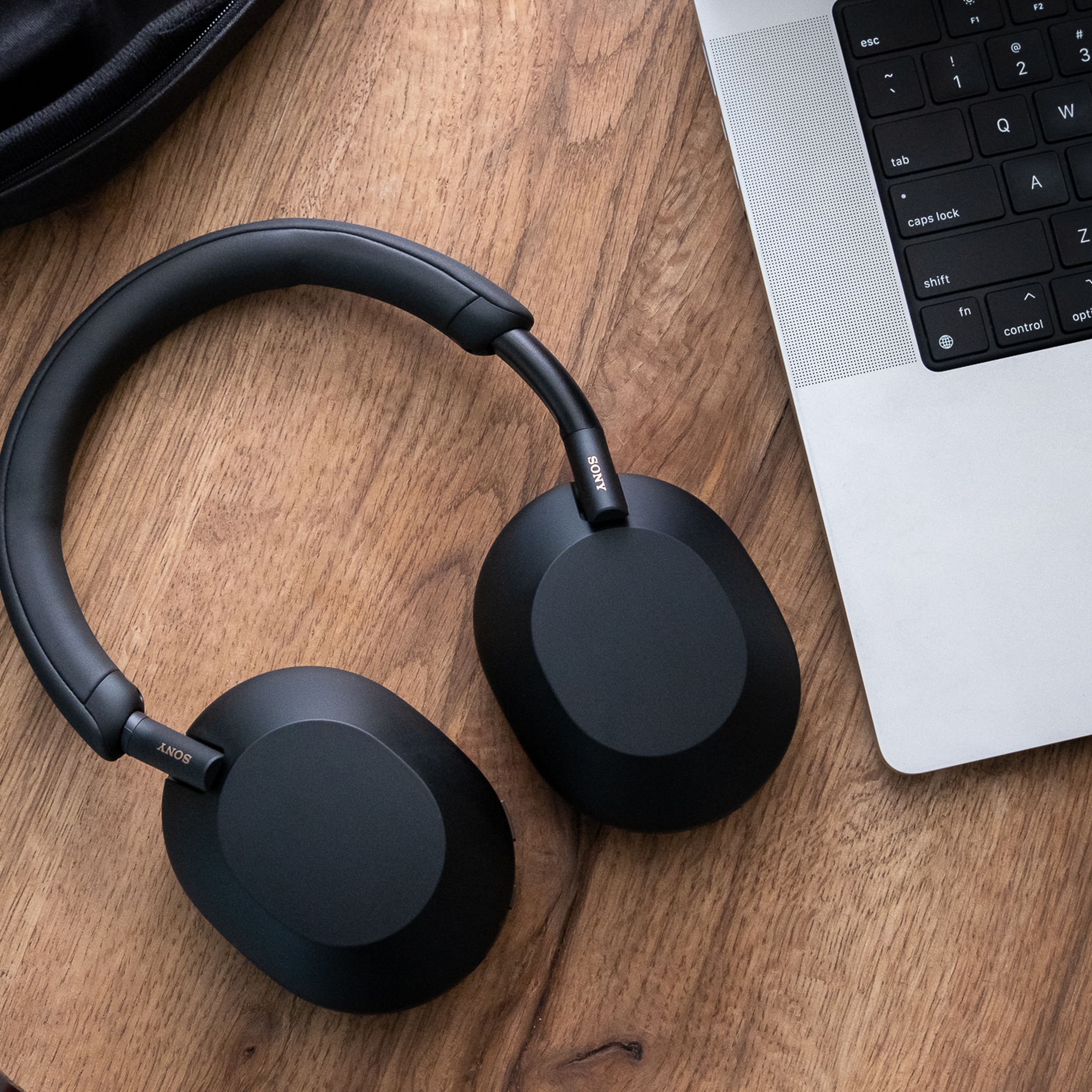 A pair of Sony WH-1000XM5 headphones, in black, resting flat on a wood table beside a laptop.